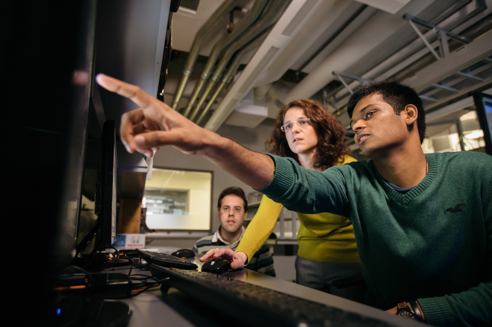student in green shirt points to computer screen while woman in yellow shirt looks on