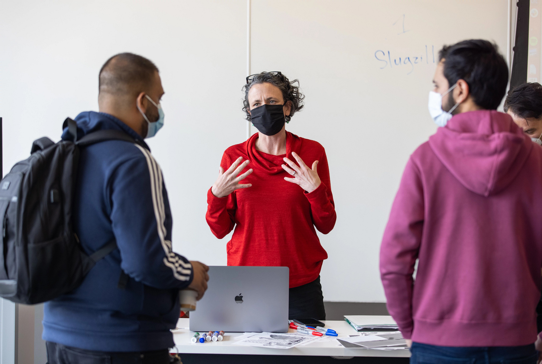 Professor in red sweater talks to two male students in front of white board