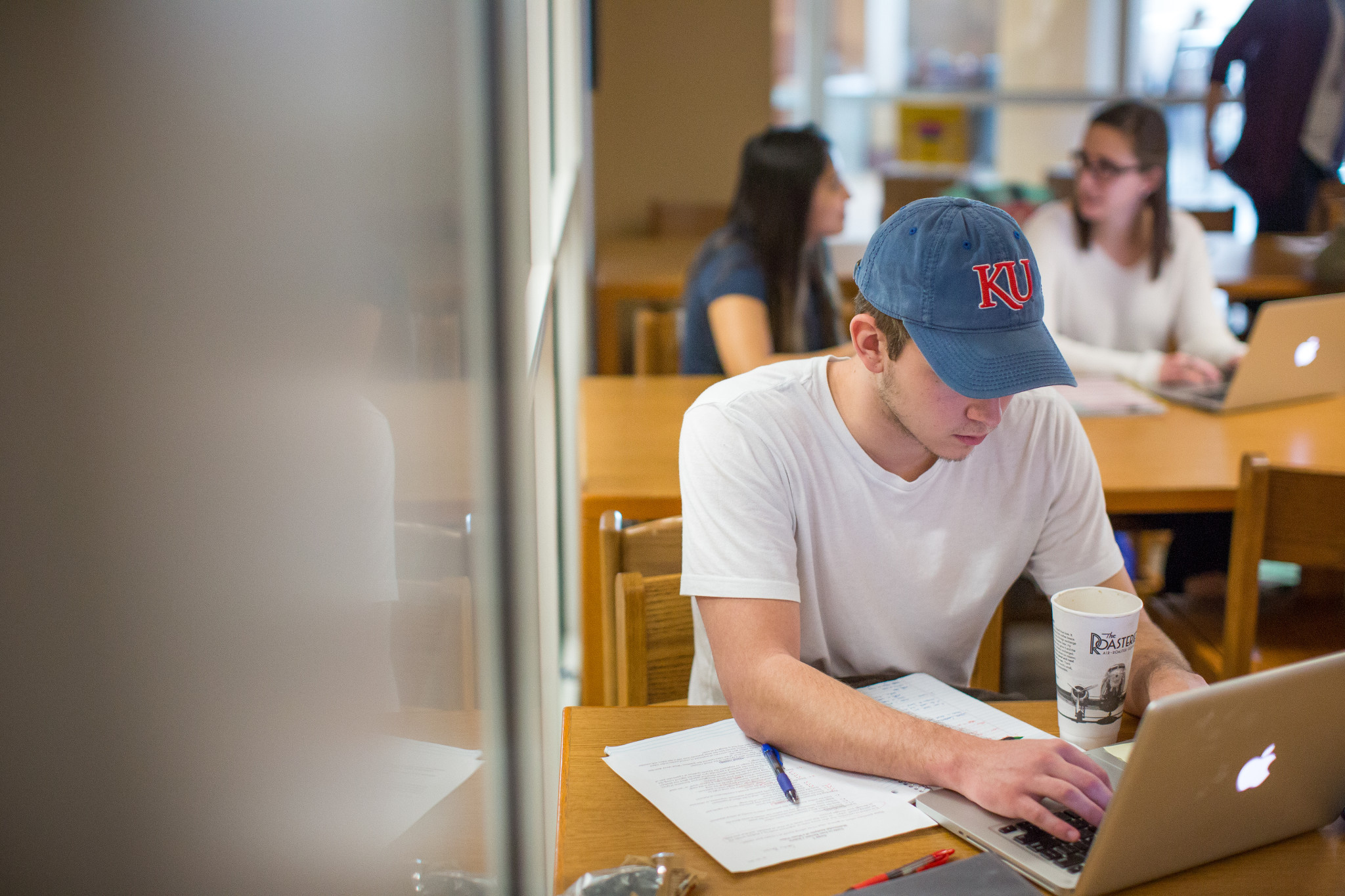 student in white t-shirt and blue hat with KU in red lettering working on a mac laptop in a library setting