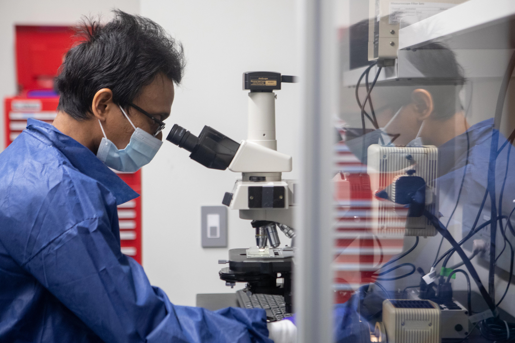 man in blue scrubs and mask looks intro microscope