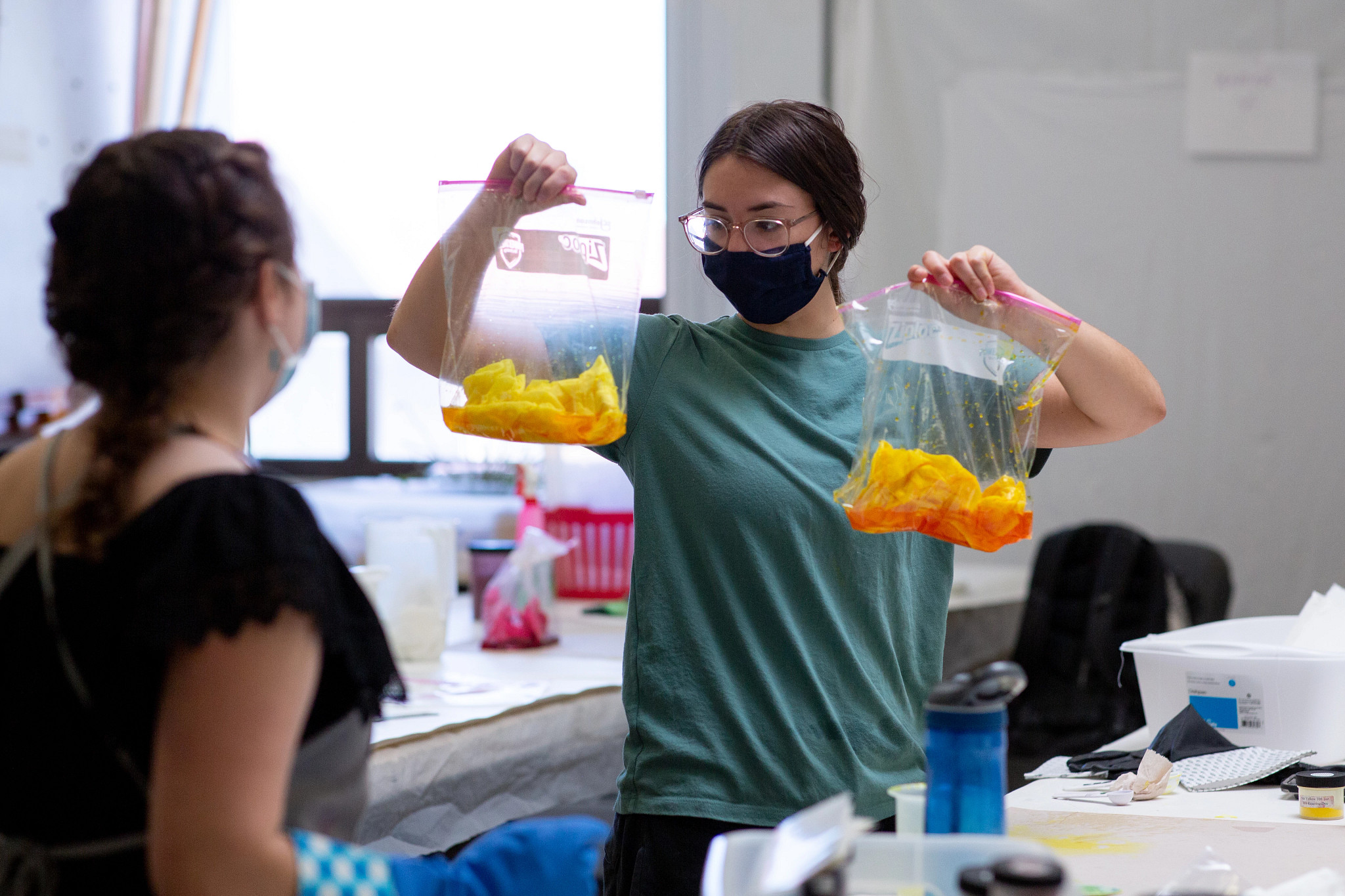 student in blue shirt showing student in black shirt two plastic bags with fabric in yellow dye