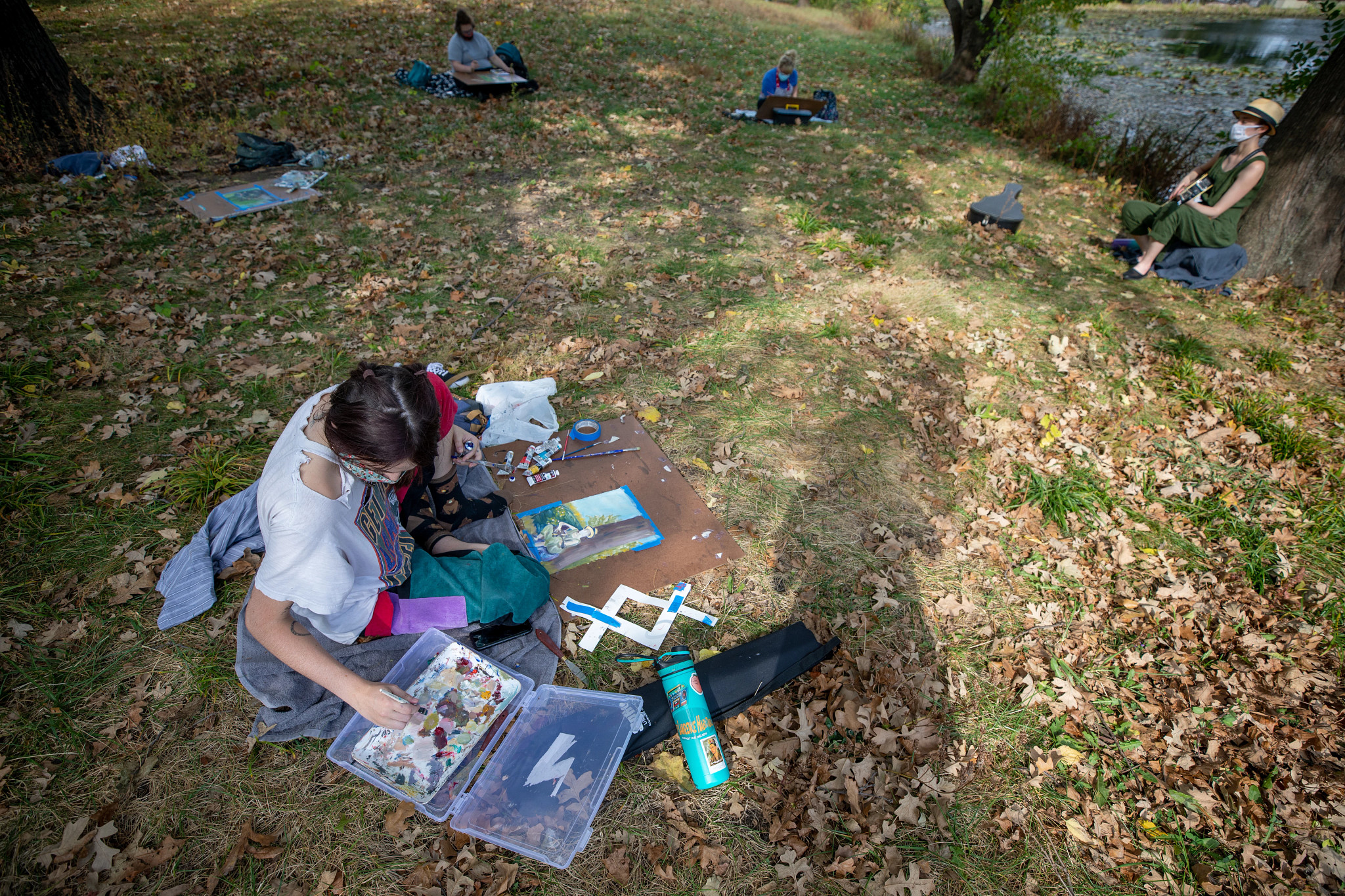 student in white shirt sits on grass with paint palette and canvas